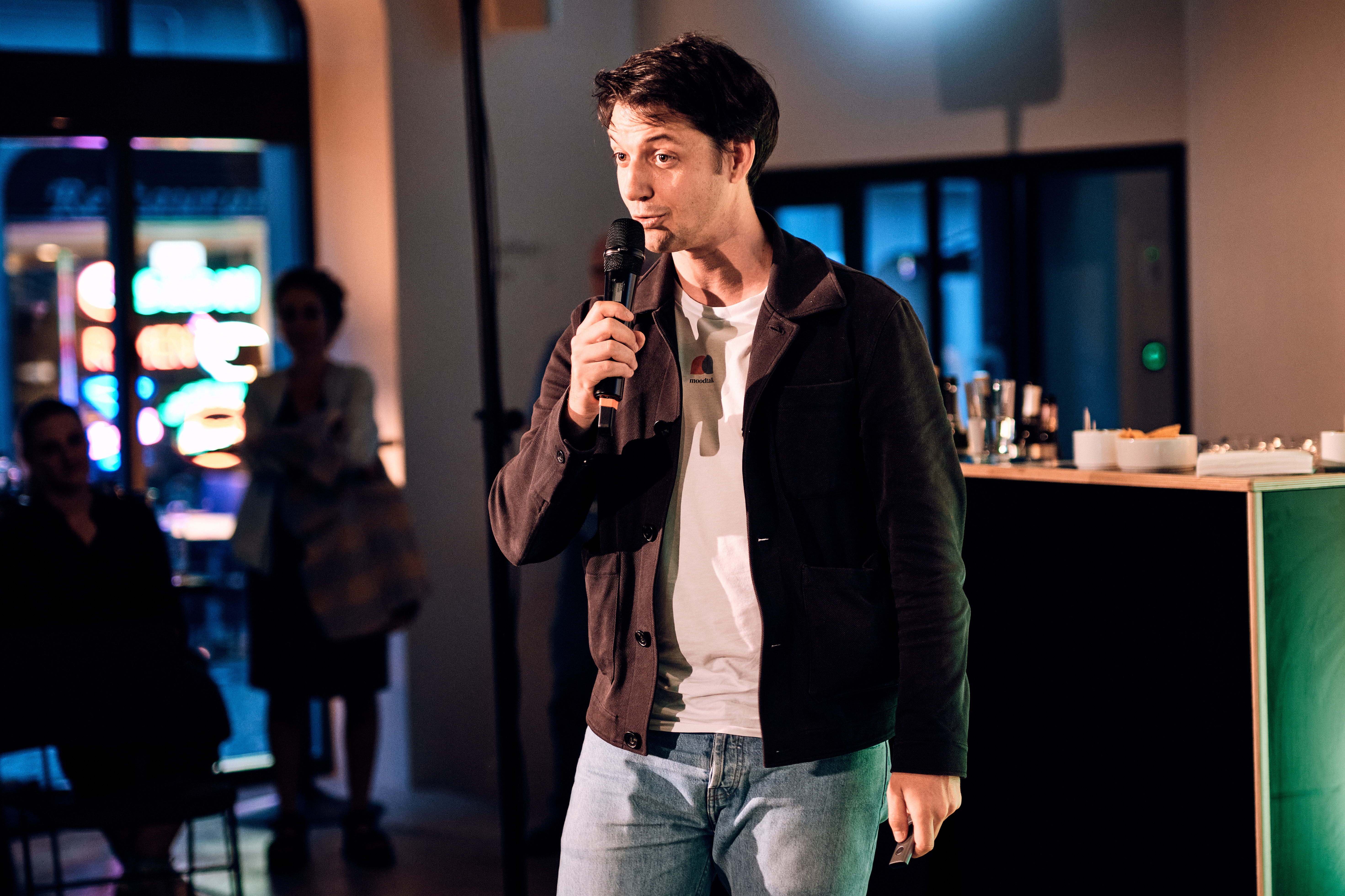 Ein Mann mit einem Mikrofon spricht in einem hell erleuchteten Innenraum, umgeben von Menschen und leuchtenden Leuchtreklamen im Hintergrund.